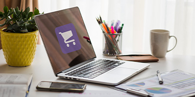 Laptop prateado em uma mesa de trabalho organizada, exibindo um ícone de carrinho de compras que simboliza a gestão eficiente de e-commerce e as soluções ERP da Alternativa Sistemas, com documentos de negócios e ferramentas de escritório ao redor, refletindo um ambiente de operações digitais.